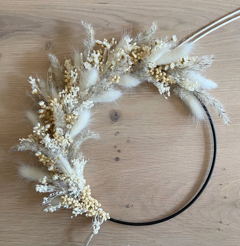 May include: A decorative wreath featuring dried flowers and grasses in shades of cream, beige, and tan. The floral arrangement is attached to a black metal ring, creating a modern, minimalist design. The wreath is perfect for adding a touch of natural beauty to any space.