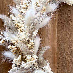 May include: Close-up of a dried floral arrangement featuring white and beige pampas grass, small white flowers, and other dried elements. The arrangement is set against a wooden background, creating a natural and textured aesthetic. Ideal for home decor.