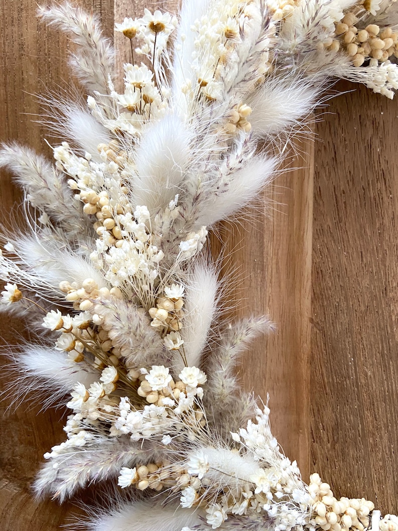 May include: Close-up of a dried floral arrangement featuring white and beige pampas grass, small white flowers, and other dried elements. The arrangement is set against a wooden background, creating a natural and textured aesthetic. Ideal for home decor.