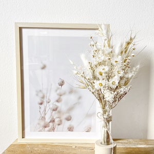 May include: A bouquet of dried white flowers in a clear glass vase with a wooden base. The vase is sitting on a wooden table in front of a framed print of a botanical illustration.