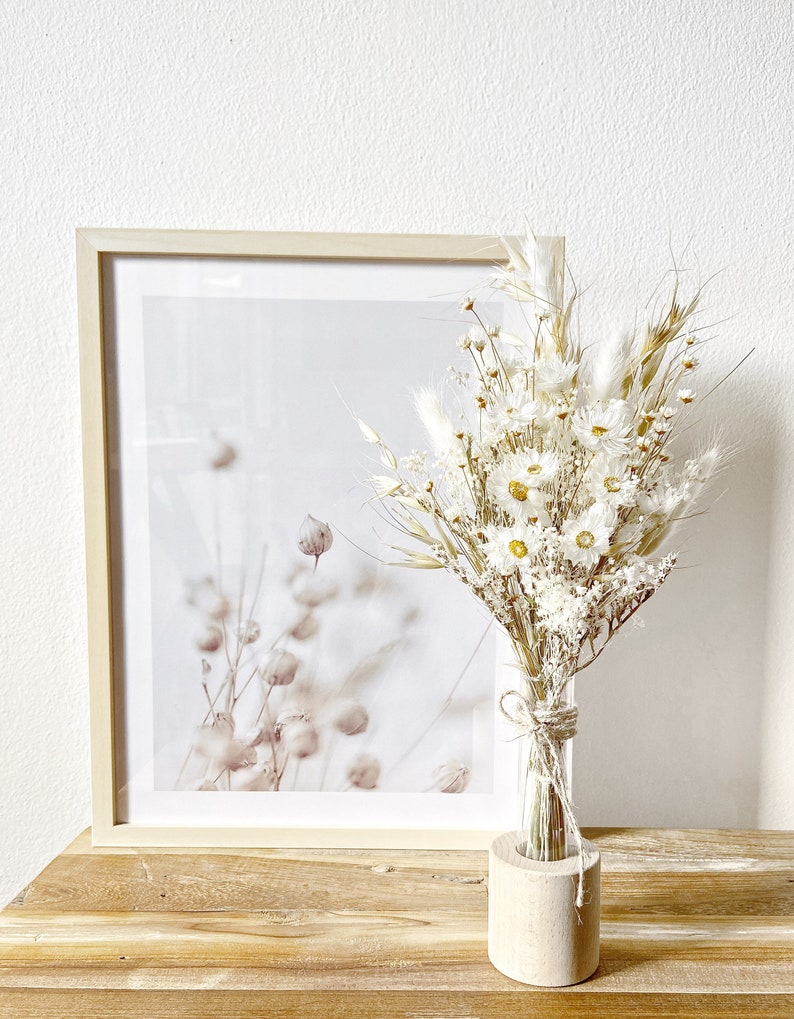 May include: A bouquet of dried white flowers in a clear glass vase with a wooden base. The vase is sitting on a wooden table in front of a framed print of a botanical illustration.