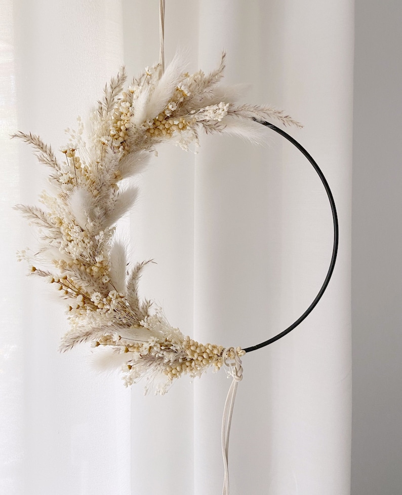 May include: A circular wall hanging featuring dried flowers and grasses in shades of cream and beige. The floral arrangement is attached to a black metal hoop, creating a modern, minimalist design. The wreath is suspended by a white cord.