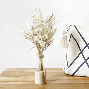 May include: A bouquet of dried white flowers in a clear glass bottle with a wooden base. The flowers are arranged in a vase on a wooden table.
