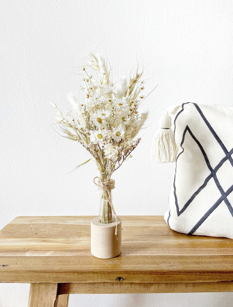 May include: A bouquet of dried white flowers in a clear glass bottle with a wooden base. The flowers are arranged in a vase on a wooden table.