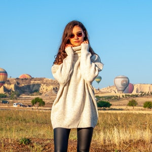 K&ouml;nnte beinhalten: Eine Frau in einem wei&szlig;en Rollkragenpullover und schwarzen Leggings steht auf einem Feld mit Hei&szlig;luftballons im Hintergrund.