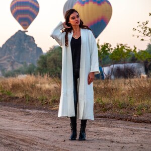 Op de afbeelding: Een vrouw in een witte, lange cardigan met lange mouwen en een zwarte broek staat in een veld met luchtballonnen op de achtergrond.
