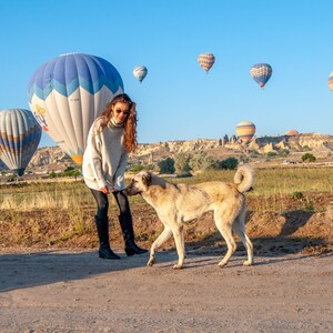 K&ouml;nnte beinhalten: Eine Frau in einem wei&szlig;en Pullover und schwarzen Hosen steht mit einem Hund auf einem Feld. Der Hund schaut die Frau an. Im Hintergrund sind Hei&szlig;luftballons zu sehen.