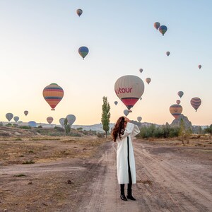 Op de afbeelding: Een vrouw in een witte jas staat op een veld met een luchtballon op de achtergrond. De ballon is rood en wit met het woord "Hotour" erop gedrukt. Er zijn veel andere luchtballonnen in de lucht.