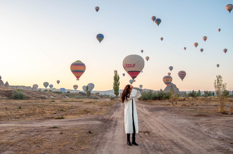 Op de afbeelding: Een vrouw in een witte jas staat op een veld met een luchtballon op de achtergrond. De ballon is rood en wit met het woord "Hotour" erop gedrukt. Er zijn veel andere luchtballonnen in de lucht.