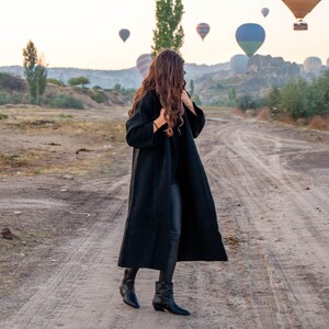 K&ouml;nnte beinhalten: Eine Frau in einem schwarzen Mantel und schwarzen Hosen steht auf einem Feldweg und blickt zu einer Gruppe von Hei&szlig;luftballons am Himmel auf.