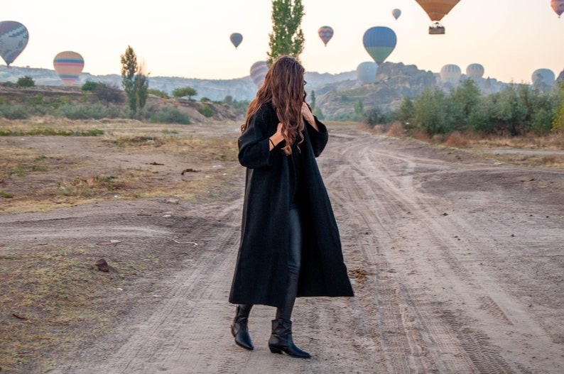 K&ouml;nnte beinhalten: Eine Frau in einem schwarzen Mantel und schwarzen Hosen steht auf einem Feldweg und blickt zu einer Gruppe von Hei&szlig;luftballons am Himmel auf.