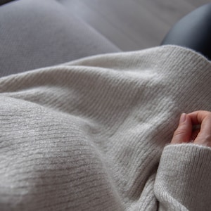 Może przedstawiać: Zbliżenie na osobę ubraną w jasnoszary sweter. Sweter ma żebrowaną fakturę i jest złożony na rękach osoby.