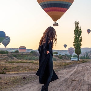K&ouml;nnte beinhalten: Eine Frau in einem schwarzen Mantel und schwarzen Hosen steht auf einem Feldweg mit einem bunten Hei&szlig;luftballon im Hintergrund. Der Ballon tr&auml;gt den Text "www.urgupballoons.com".