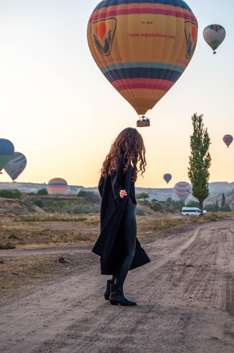 K&ouml;nnte beinhalten: Eine Frau in einem schwarzen Mantel und schwarzen Hosen steht auf einem Feldweg mit einem bunten Hei&szlig;luftballon im Hintergrund. Der Ballon tr&auml;gt den Text "www.urgupballoons.com".