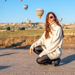 K&ouml;nnte beinhalten: Eine Frau in einem wei&szlig;en Rollkragenpullover und schwarzen Leggings posiert vor einem Feld mit Hei&szlig;luftballons im Hintergrund.