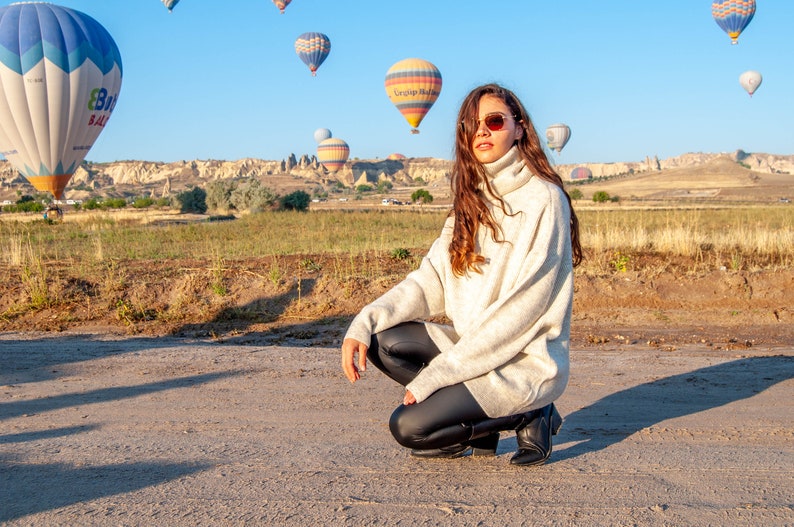 K&ouml;nnte beinhalten: Eine Frau in einem wei&szlig;en Rollkragenpullover und schwarzen Leggings posiert vor einem Feld mit Hei&szlig;luftballons im Hintergrund.