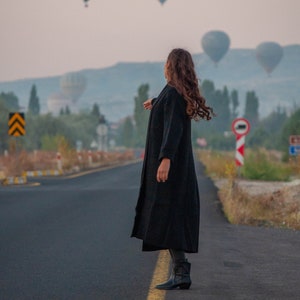 K&ouml;nnte beinhalten: Eine Person in einem schwarzen Mantel und schwarzen Stiefeln steht auf einer asphaltierten Stra&szlig;e und blickt zu Hei&szlig;luftballons am Himmel auf.