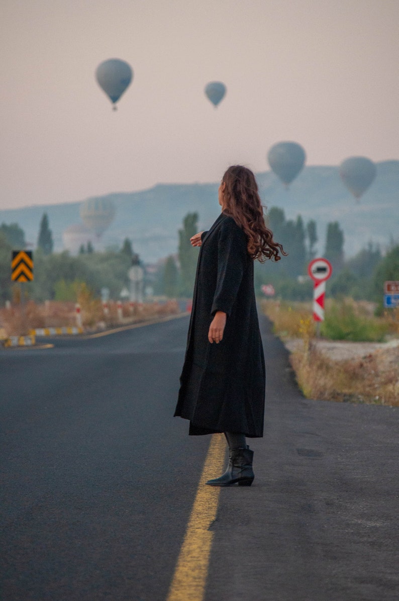 K&ouml;nnte beinhalten: Eine Person in einem schwarzen Mantel und schwarzen Stiefeln steht auf einer asphaltierten Stra&szlig;e und blickt zu Hei&szlig;luftballons am Himmel auf.