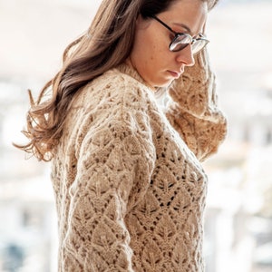 May include: A close-up of a woman wearing a beige knitted sweater with a delicate pattern.