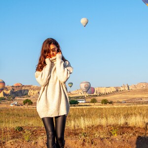 K&ouml;nnte beinhalten: Eine Frau in einem wei&szlig;en Rollkragenpullover und schwarzen Leggings steht auf einem Feld mit Hei&szlig;luftballons im Hintergrund. Die Ballons sind in verschiedenen Farben und Formen und einige haben Logos.