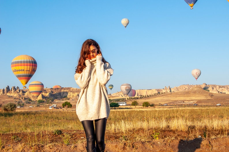 K&ouml;nnte beinhalten: Eine Frau in einem wei&szlig;en Rollkragenpullover und schwarzen Leggings steht auf einem Feld mit Hei&szlig;luftballons im Hintergrund. Die Ballons sind in verschiedenen Farben und Formen und einige haben Logos.
