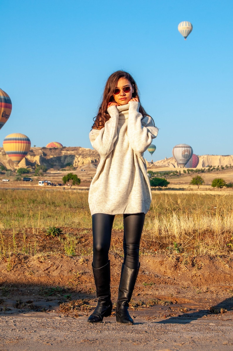 K&ouml;nnte beinhalten: Eine Frau in einem wei&szlig;en Rollkragenpullover und schwarzen Leggings steht auf einem Feld mit Hei&szlig;luftballons im Hintergrund.