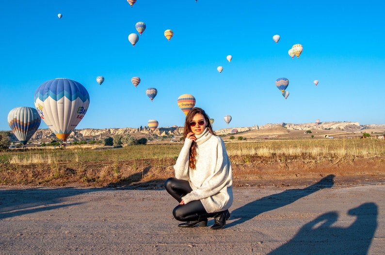 K&ouml;nnte beinhalten: Eine Frau in einem wei&szlig;en Rollkragenpullover und schwarzen Leggings posiert vor einem Feld mit Hei&szlig;luftballons. Die Ballons sind in verschiedenen Farben und Formen, und der Himmel ist blau.