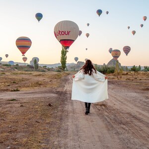 Op de afbeelding: Een vrouw in een witte jas loopt over een zandpad met een veld vol luchtballonnen op de achtergrond. De ballonnen zijn in verschillende kleuren en sommige hebben logo's.