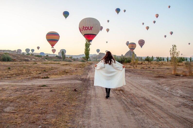 Op de afbeelding: Een vrouw in een witte jas loopt over een zandpad met een veld vol luchtballonnen op de achtergrond. De ballonnen zijn in verschillende kleuren en sommige hebben logo's.