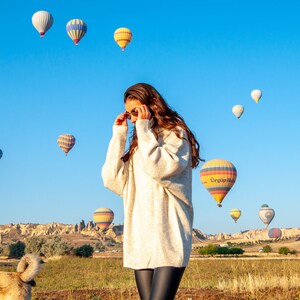 K&ouml;nnte beinhalten: Eine Frau in einem wei&szlig;en Pullover und schwarzen Leggings steht auf einem Feld mit einer Gruppe von Hei&szlig;luftballons am Himmel. Die Ballons sind in verschiedenen Farben und Formen und einige haben Logos. Die Frau schaut zu den Ballons hinauf und der Himmel ist blau.