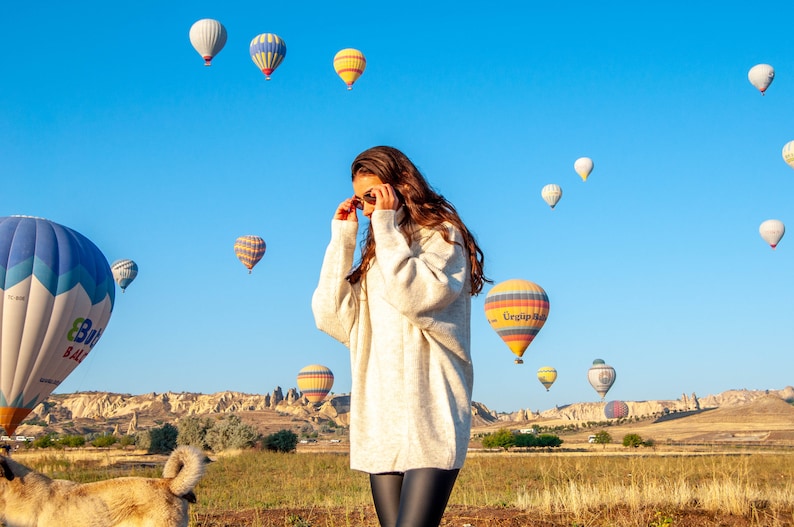 K&ouml;nnte beinhalten: Eine Frau in einem wei&szlig;en Pullover und schwarzen Leggings steht auf einem Feld mit einer Gruppe von Hei&szlig;luftballons am Himmel. Die Ballons sind in verschiedenen Farben und Formen und einige haben Logos. Die Frau schaut zu den Ballons hinauf und der Himmel ist blau.