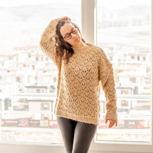 May include: A woman wearing a beige knitted sweater and black pants.