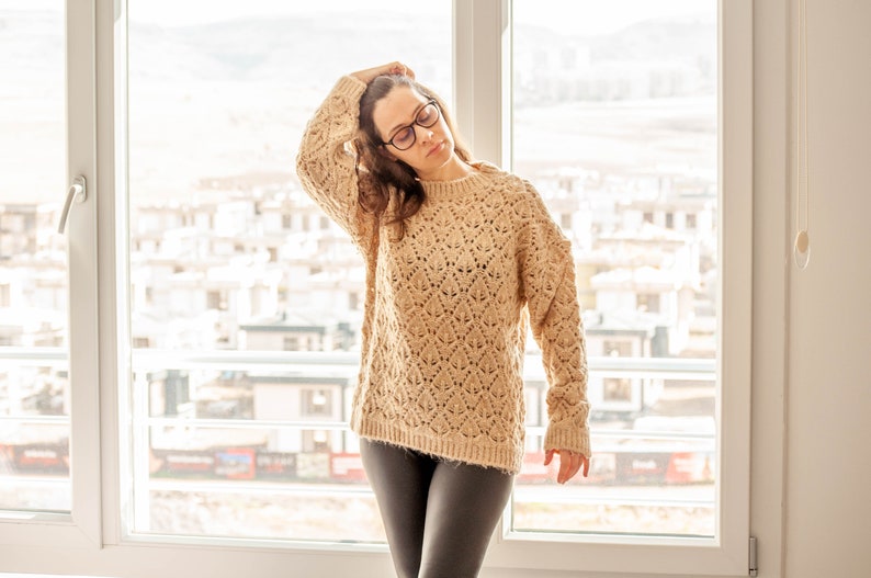 May include: A woman wearing a beige knitted sweater and black pants.
