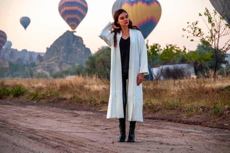 Op de afbeelding: Een vrouw in een witte, lange cardigan met lange mouwen en een zwarte broek staat in een veld met luchtballonnen op de achtergrond.