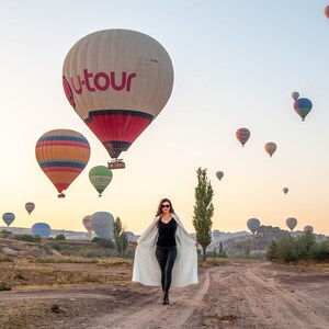 Op de afbeelding: Een vrouw in een witte jas loopt in een veld richting de camera met uitzicht op luchtballonnen in de lucht. De ballonnen zijn in verschillende kleuren en maten, met &eacute;&eacute;n grote ballon met het woord "Utour" in rode letters.