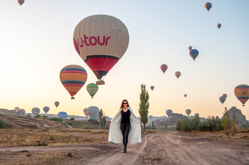 Op de afbeelding: Een vrouw in een witte jas loopt in een veld richting de camera met uitzicht op luchtballonnen in de lucht. De ballonnen zijn in verschillende kleuren en maten, met &eacute;&eacute;n grote ballon met het woord "Utour" in rode letters.