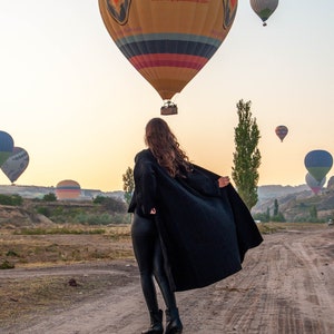 K&ouml;nnte beinhalten: Eine Frau in einem schwarzen Mantel und schwarzen Hosen steht auf einem Feldweg und blickt zu einem Hei&szlig;luftballon auf. Der Ballon ist gelb, rot und blau mit dem Text "www.urgupballoons.com" und "TC-BUK".