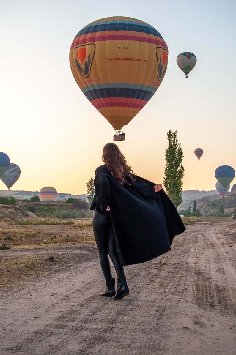 K&ouml;nnte beinhalten: Eine Frau in einem schwarzen Mantel und schwarzen Hosen steht auf einem Feldweg und blickt zu einem Hei&szlig;luftballon auf. Der Ballon ist gelb, rot und blau mit dem Text "www.urgupballoons.com" und "TC-BUK".