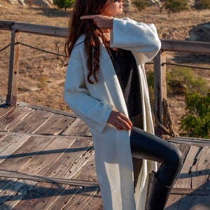 Op de afbeelding: Een vrouw draagt een witte lange mouwen cardigan, zwarte leggings en zwarte laarzen. Ze staat op een houten platform met uitzicht op een rotsachtig landschap op de achtergrond.