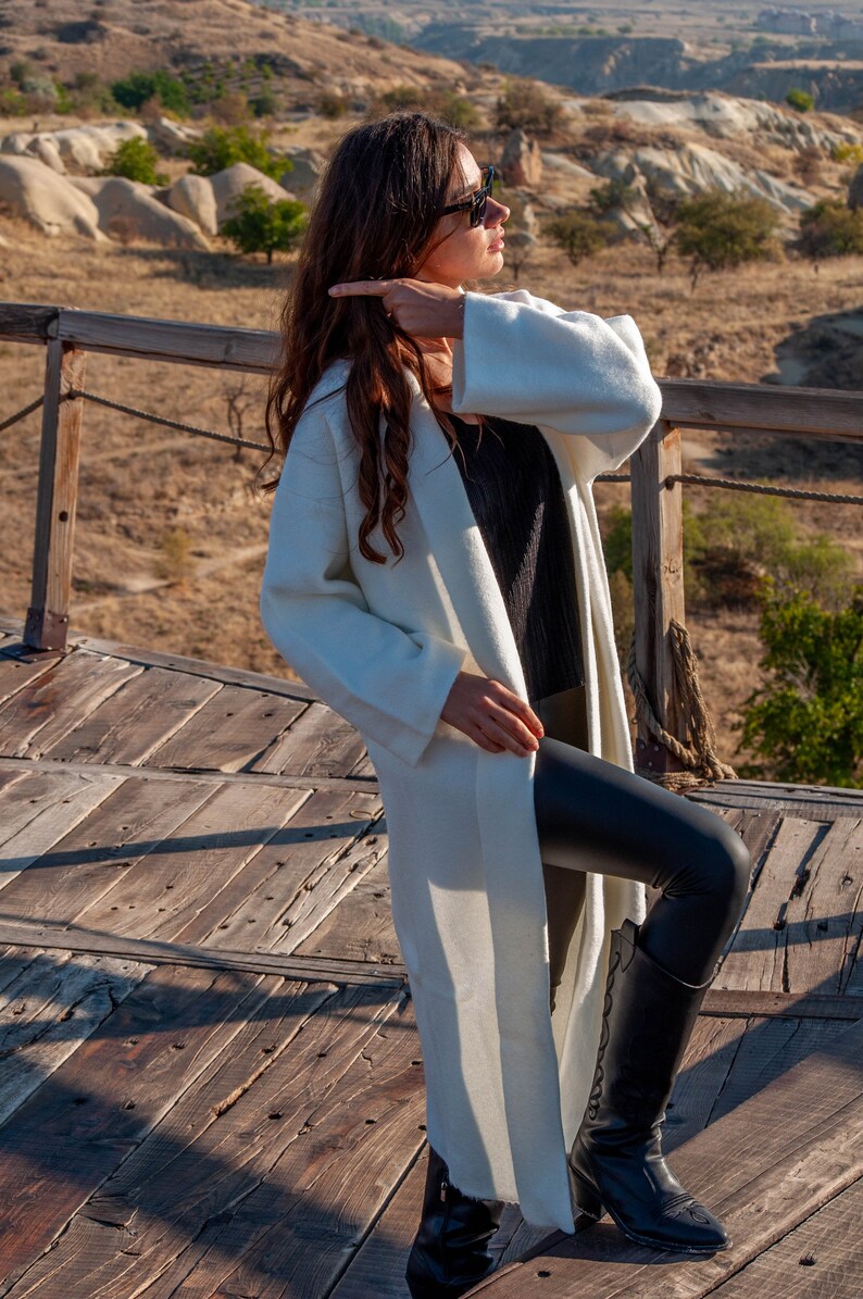Op de afbeelding: Een vrouw draagt een witte lange mouwen cardigan, zwarte leggings en zwarte laarzen. Ze staat op een houten platform met uitzicht op een rotsachtig landschap op de achtergrond.