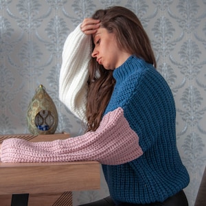 May include: A woman wearing a blue, pink and white knitted sweater with long sleeves. She is sitting at a table with her head in her hand.