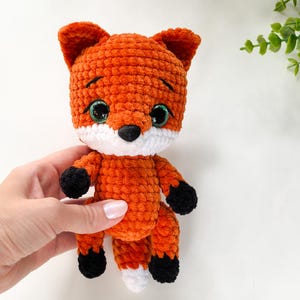 Op de afbeelding: Een gehaakt oranje vossen knuffel met zwarte ogen, een witte neus en zwarte poten. De vos zit op een wit oppervlak.