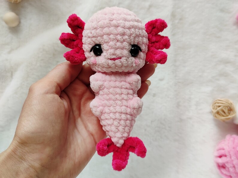 May include: A pink and red crocheted axolotl plush toy. The axolotl has black eyes and a pink body with red gills and tail.