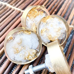 May include: A set of three round cabinet knobs featuring a clear resin inlay with embedded gold leaf flakes. Each knob is encased in a circular brass-toned metal frame and includes a threaded metal bolt and nut for secure installation on furniture.