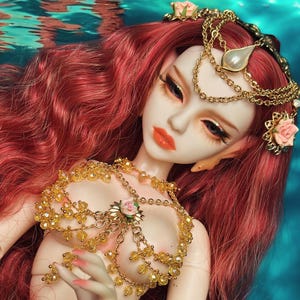 Peut inclure: Une poupée aux longs cheveux roux et un diadème doré avec une perle. La poupée porte un haut doré orné de perles et une jupe rouge avec une ceinture dorée. Des roses roses ornent les accessoires de la poupée. La poupée est immergée dans l'eau bleue.