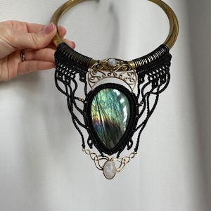 May include: A statement necklace featuring a large, iridescent labradorite stone pendant. The necklace has a brass-colored collar, black macrame detailing, and small moonstone accents. The design is bohemian and handcrafted.