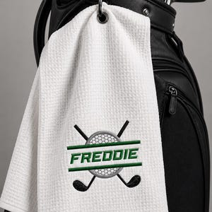 May include: A white waffle-textured golf towel featuring an embroidered design of a golf ball and two crossed golf clubs. The name FREDDIE is stitched in green capital letters across the centre of the golf ball graphic on the fabric.