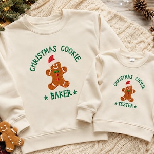 May include: Two cream-colored crewneck sweatshirts featuring embroidered gingerbread men wearing Santa hats. The larger sweatshirt reads "CHRISTMAS COOKIE BAKER" in green text, while the smaller one reads "CHRISTMAS COOKIE TESTER" in green text.