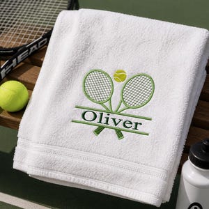 Könnte beinhalten: Ein weißes Baumwollhandtuch mit einer Stickerei aus zwei gekreuzten grünen Tennisschlägern und einem gelben Tennisball. Der Name Oliver ist in dunkelgrünen Buchstaben auf einer horizontalen Linie über den Schlägergriffen eingestickt.