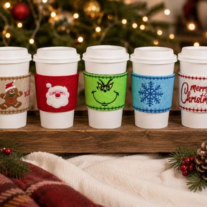 Op de afbeelding: Vijf witte wegwerp koffiebekers met vilten hoezen. De hoezen zijn voorzien van een peperkoekmannetje, een kerstmangezicht, een groen karaktergezicht, een blauwe sneeuwvlok en de tekst Merry Christmas met een hulsttakje.